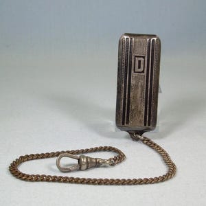 May include: A silver-toned pocket watch case with a rectangular shape, vertical lines, and a central capital "D". It is connected to a chain with a clasp. The item is a vintage pocket watch case.