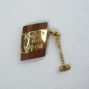 May include: A vintage "Knock on Wood" charm. The charm features a wooden base with a gold-colored metal plaque that reads "Knock on Wood". A small gold-colored gavel hangs from the charm.