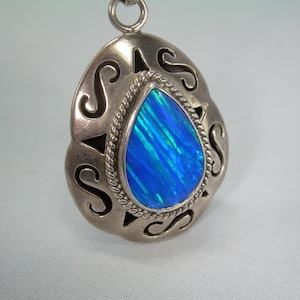May include: A teardrop-shaped pendant featuring a central blue iridescent stone set within a twisted metal border. The surrounding silver-toned metal frame has a smooth finish with cut-out S-shaped patterns and a small circular bail at the top.