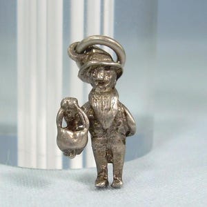 Puede incluir: Un dije de metal en tono plateado que representa una figura con un sombrero de ala ancha y barba puntiaguda. La figura sostiene un objeto pequeño y redondeado en una mano y cuenta con una argolla circular en la parte superior del sombrero.
