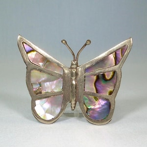 May include: A silver-toned butterfly brooch with iridescent abalone shell inlay. The butterfly has detailed wings and antennae. The shell pieces reflect pink, purple, and green hues, creating a shimmering effect. A decorative jewelry item.