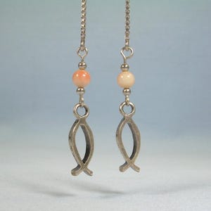 May include: A pair of dangle earrings featuring a silver fish symbol and a peach-colored bead. The earrings hang from a delicate silver chain. The fish symbol is a classic religious emblem.