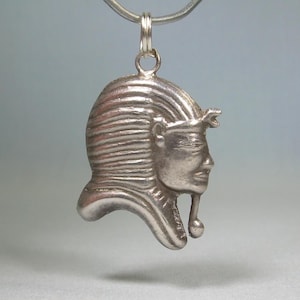Op de afbeelding: Een zilveren hanger met het profiel van een Egyptische farao. De hanger heeft gedetailleerde gelaatstrekken, een gestreepte hoofdtooi en een kleine lus voor een ketting. De hanger hangt aan een zilveren ketting.