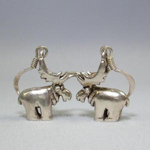 May include: A pair of silver-toned moose-shaped earrings with curved ear wires. Each earring features a detailed moose figure with prominent antlers and a smooth, polished finish. The metal is stamped with 925 to indicate sterling silver composition.