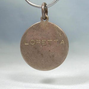 May include: A round, silver-toned pendant with the name "LORETTA" engraved in capital letters. The pendant is suspended from a silver chain. The background is a soft, blurred gradient of blue and white.