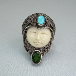 May include: A silver ring featuring a carved ivory-colored face with a turquoise stone above and a green stone below. The ring has intricate detailing around the face and stones, with a textured band.