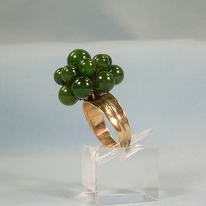 May include: A gold ring set with a cluster of dark green jade stones. The ring is displayed on a clear acrylic stand.