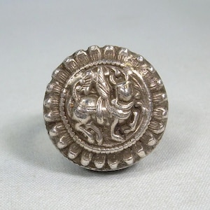 May include: A silver-colored, round button with an embossed design. The central image features a horse and rider within a decorative border. The button has a scalloped edge and is set against a light background.
