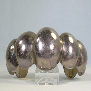 May include: A silver bracelet featuring a series of connected, oval links. The bracelet is displayed on a clear stand against a light blue backdrop. The metal has a polished, reflective surface.
