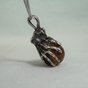 May include: A silver pendant featuring a detailed skeletal hand holding a round amber-colored gemstone.