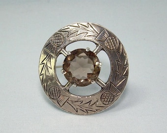 May include: A circular silver brooch featuring an engraved thistle pattern around the perimeter and a central, round, faceted brown gemstone held in place by four prongs. The metal has a polished finish with dark recessed areas in the engraving.