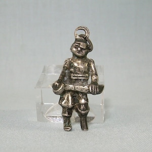 May include: A silver charm depicting a small figure of a soldier holding a weapon. The charm has a loop at the top for attaching to a bracelet or necklace.