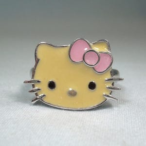 May include: A silver ring featuring a yellow enamel face with black eyes and whiskers. The ring has a pink bow and is a popular character design. The ring is a cute accessory.