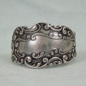 Può includere: Un anello largo color argento con un design ornamentale a volute. L'anello presenta un'area centrale rettangolare con una superficie liscia, circondata da motivi a spirale dettagliati. Il design dell'anello è simmetrico e ha un'estetica vintage.