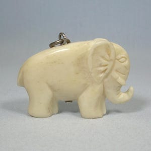 Puede incluir: Figurita de elefante de color marfil con una trompa, orejas y ojos detallados. La pequeña escultura tiene un anillo de metal adjunto en la parte superior. Este elefante decorativo es un coleccionable encantador.