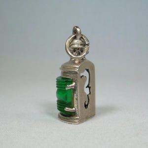 May include: A silver-coloured lantern charm with a bright green rectangular jewel. The charm has a detailed top with a loop for attachment and a cutout design on the side. The charm is approximately 2.5 cm in height.