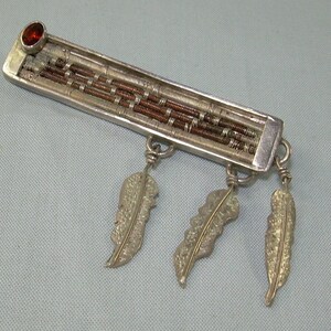 May include: A silver pendant with a red gemstone and three silver feather charms. The pendant is made of woven silver and copper wire.