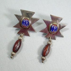 May include: A pair of silver-tone earrings. Each earring features a starburst design with a blue and red center. Below the starburst hangs a dark red bead with white stripes and silver accents. The earrings are displayed on a light background.