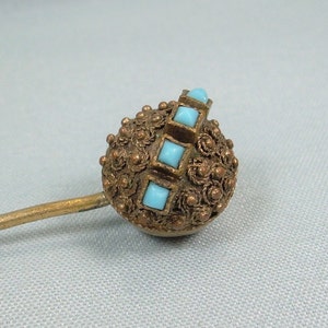 Op de afbeelding: Een gouden stickpin met een ronde, sierlijke kop. De kop is versierd met een herhalend patroon van kleine, verhoogde cirkels en een rij vierkante turquoise blauwe stenen.