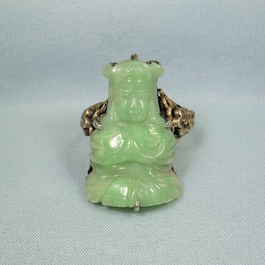 May include: A jade green ring with a carved figure of a person. The ring is made of silver and has a detailed design.