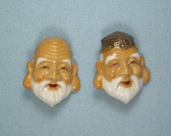 May include: Two ceramic figurines of smiling, bearded men with large ears. One figurine is wearing a brown hat with gold accents. Both figurines have white beards and brown skin.