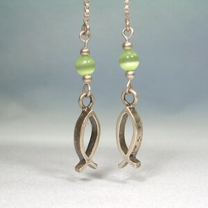 May include: Silver earrings featuring a fish symbol pendant. Each earring has a light green bead and a small silver chain. The fish pendants have a textured, antique finish. The earrings are displayed against a soft, blurred background.