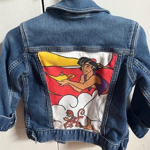 May include: A blue denim jacket featuring a vibrant graphic of Aladdin, a genie lamp, and Abu the monkey on the back. The jacket has a classic collar, button closures, and long sleeves.