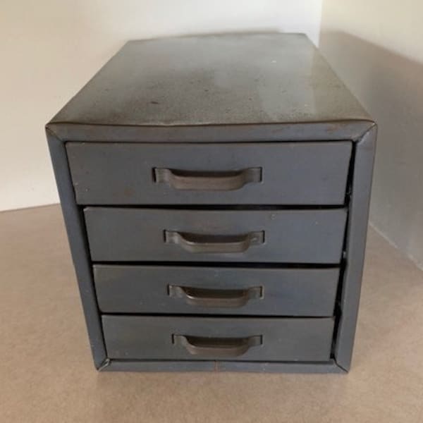 May include: A small, gray metal cabinet with four drawers. Each drawer has a curved handle. The cabinet is rectangular and sits on a light-colored surface. The cabinet is a muted gray color.