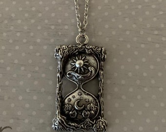May include: A silver necklace with an hourglass pendant featuring a sun and moon design. The pendant is detailed with stars and clouds.