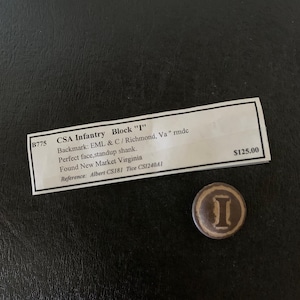 May include: A brown and tan Civil War era button with the letter "I" in the center. The button is on a black surface next to a white label with text that reads "CSA Infantry Block 'I'" and other details.