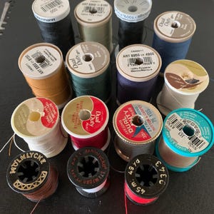 May include: A collection of fourteen assorted sewing thread spools in various colors including black, white, red, blue, green, brown, and tan. The spools feature different brands such as Dual Duty, Coats & Clark, and Molnlycke with visible labels.