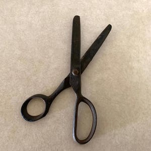 May include: A pair of vintage scissors with dark metal blades and finger holes. The scissors are open, displaying the blades and the pivot point. The handles have a rounded design.
