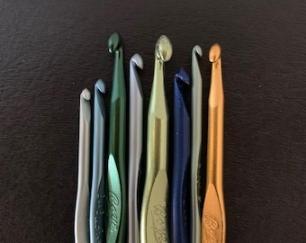 May include: A collection of crochet hooks in various metallic colors, including silver, green, gold, and blue. The hooks are arranged vertically, showcasing their different sizes and designs. The hooks have the word "Bates" engraved on them.