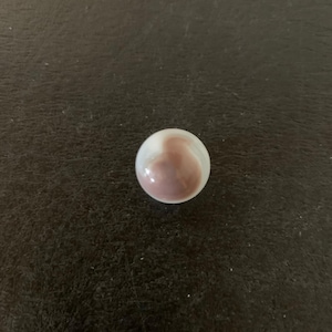 May include: A single marble with a white base and a swirling pattern of mauve and light brown. The marble is round and smooth, with a glossy finish. The background is a dark, textured surface.