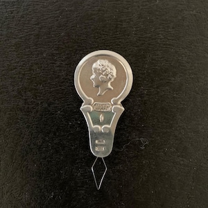 May include: A silver-toned needle threader with a circular top featuring a profile portrait. The threader has a decorative handle and a diamond-shaped wire loop for threading needles. The word "JAPAN" is visible on the handle.