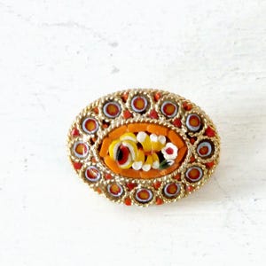 May include: An oval-shaped brooch with a floral design made of colorful mosaic tiles. The brooch is surrounded by a gold-colored frame with small circles.