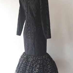 May include: Black vintage dress with long sleeves and a V-neck. The bodice has a textured pattern, and the skirt features a tulle overlay with gold circle accents. The dress is displayed on a mannequin.