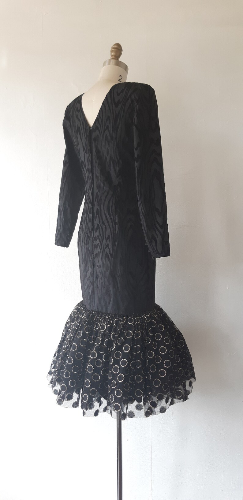 May include: Black vintage dress with long sleeves and a V-neck. The bodice has a textured pattern, and the skirt features a tulle overlay with gold circle accents. The dress is displayed on a mannequin.