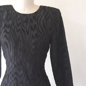 May include: A black, long-sleeved dress with a unique, textured pattern. The dress is displayed on a mannequin, showcasing its form-fitting silhouette and round neckline. The fabric appears to have a subtle sheen.