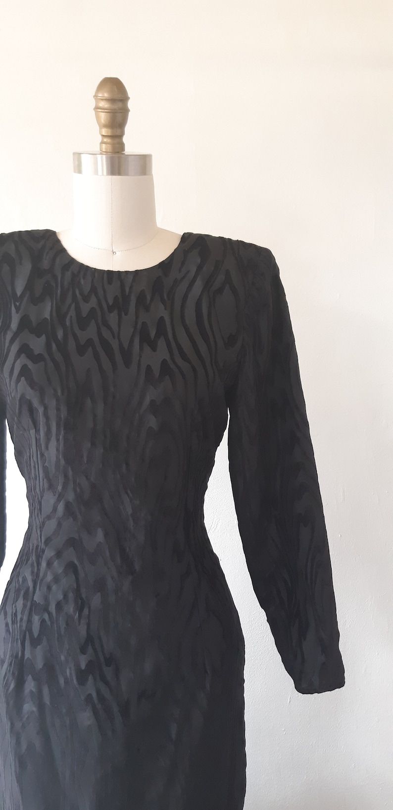 May include: A black, long-sleeved dress with a unique, textured pattern. The dress is displayed on a mannequin, showcasing its form-fitting silhouette and round neckline. The fabric appears to have a subtle sheen.