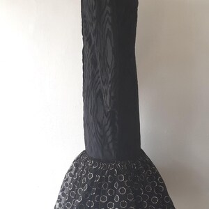 May include: A black formal dress with a textured bodice and a flared skirt. The skirt features a sheer overlay with a pattern of gold-colored circles. The dress is displayed on a mannequin.