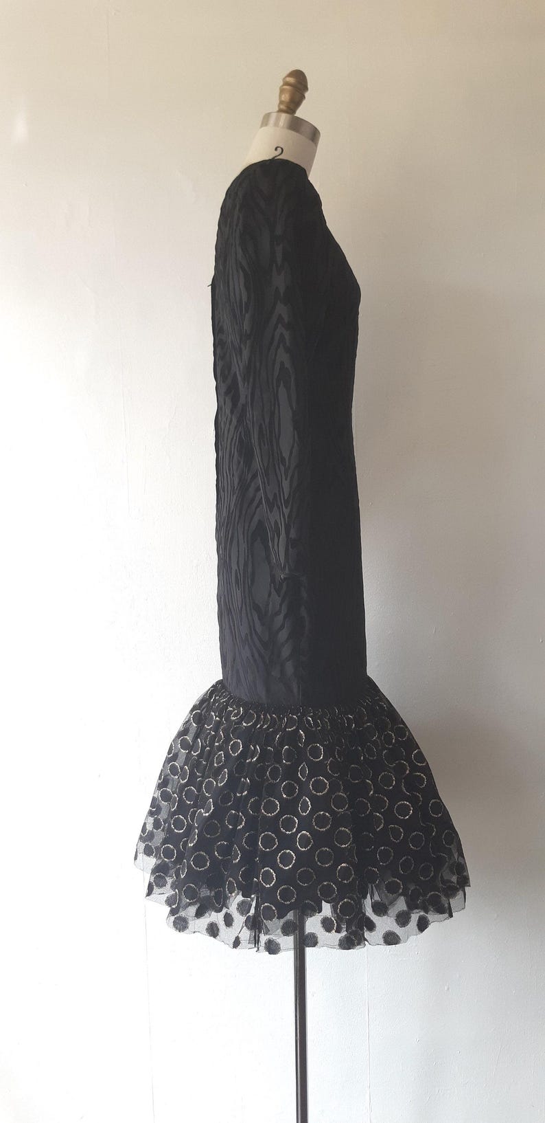 May include: A black formal dress with a textured bodice and a flared skirt. The skirt features a sheer overlay with a pattern of gold-colored circles. The dress is displayed on a mannequin.