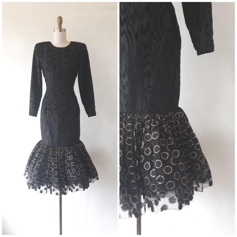 May include: Black long-sleeve dress with a textured pattern. The dress features a fitted bodice and a flared skirt with a sheer overlay adorned with gold-colored circles. The dress is displayed on a mannequin.