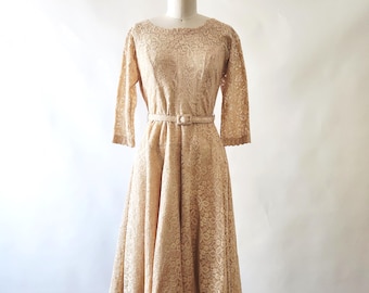 May include: A beige lace dress featuring a round neckline, three-quarter length sleeves, and a flared skirt. The dress includes a matching thin belt with a rectangular buckle at the waist, showcasing an intricate floral pattern throughout the fabric.