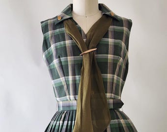 May include: A sleeveless dress featuring a green, white, and grey plaid pattern with a collared neckline and gold-tone buttons. The dress includes a matching fabric belt at the waist and is accented with a sheer olive green scarf secured by a gold pin.