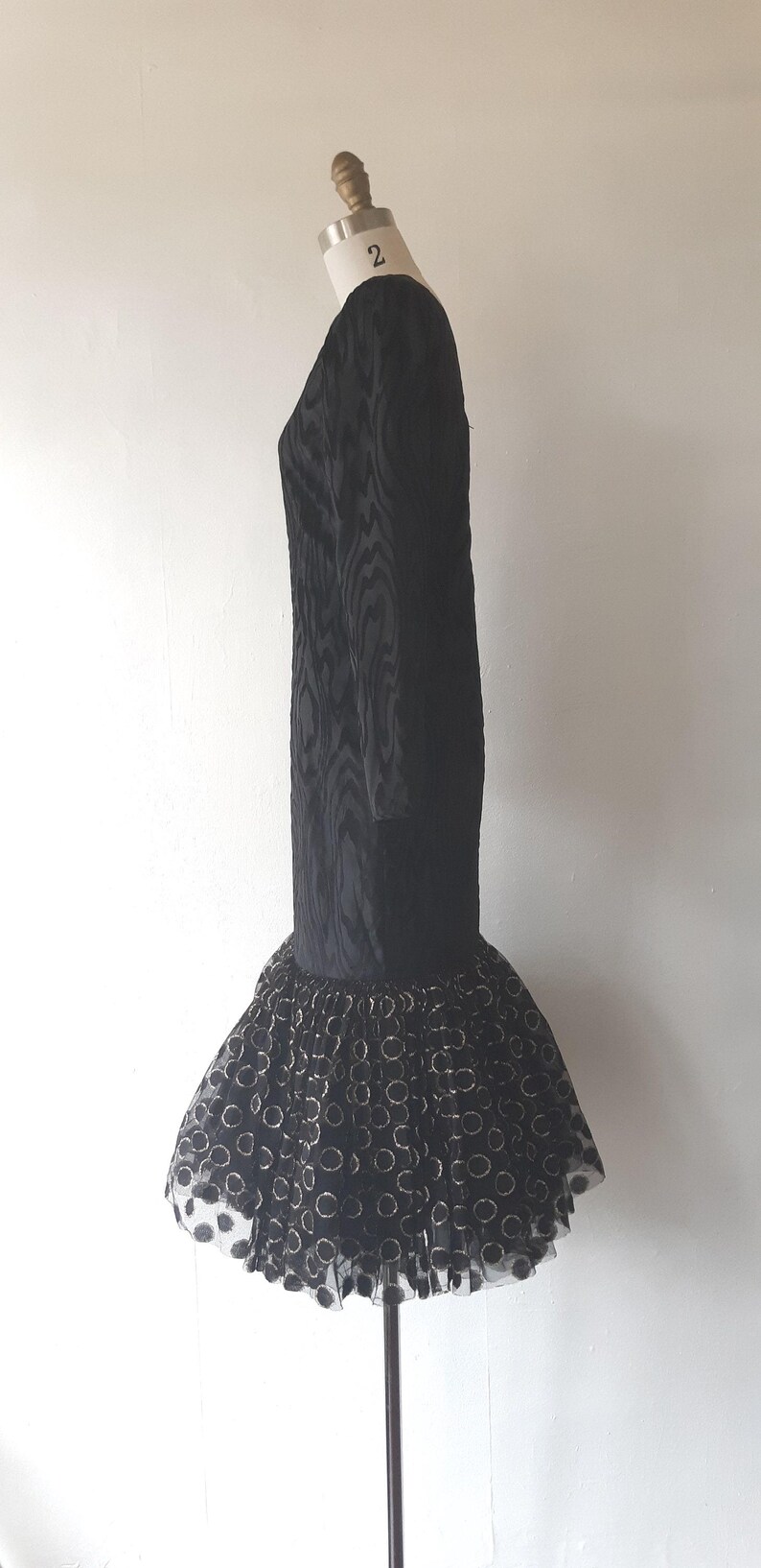 May include: A black dress with long sleeves and a textured pattern. The dress has a flared skirt with a pattern of gold circles on a black tulle base. The dress is displayed on a mannequin.