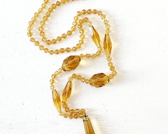 Peut inclure: Un collier vintage de couleur ambre avec une variété de perles à facettes et un pendentif allongé. Le collier comporte de petites perles rondes entrecoupées de perles plus grandes, de forme ovale. Le pendentif est un long cristal pointu. Le collier est présenté sur une surface blanche.