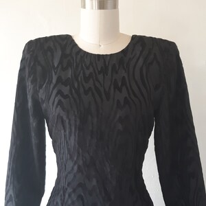 May include: A black, long-sleeved dress with a unique zebra-like pattern. The dress is displayed on a mannequin, showcasing its fitted silhouette and round neckline. The fabric has a subtle sheen.