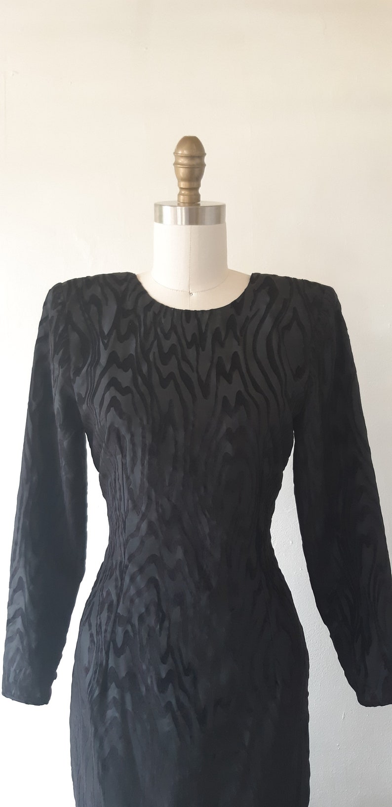 May include: A black, long-sleeved dress with a unique zebra-like pattern. The dress is displayed on a mannequin, showcasing its fitted silhouette and round neckline. The fabric has a subtle sheen.