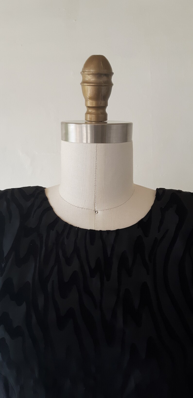 May include: A black top with a velvet zebra stripe pattern is displayed on a mannequin. The mannequin has a cream-colored neck and a gold-colored head. The top has a scoop neckline.